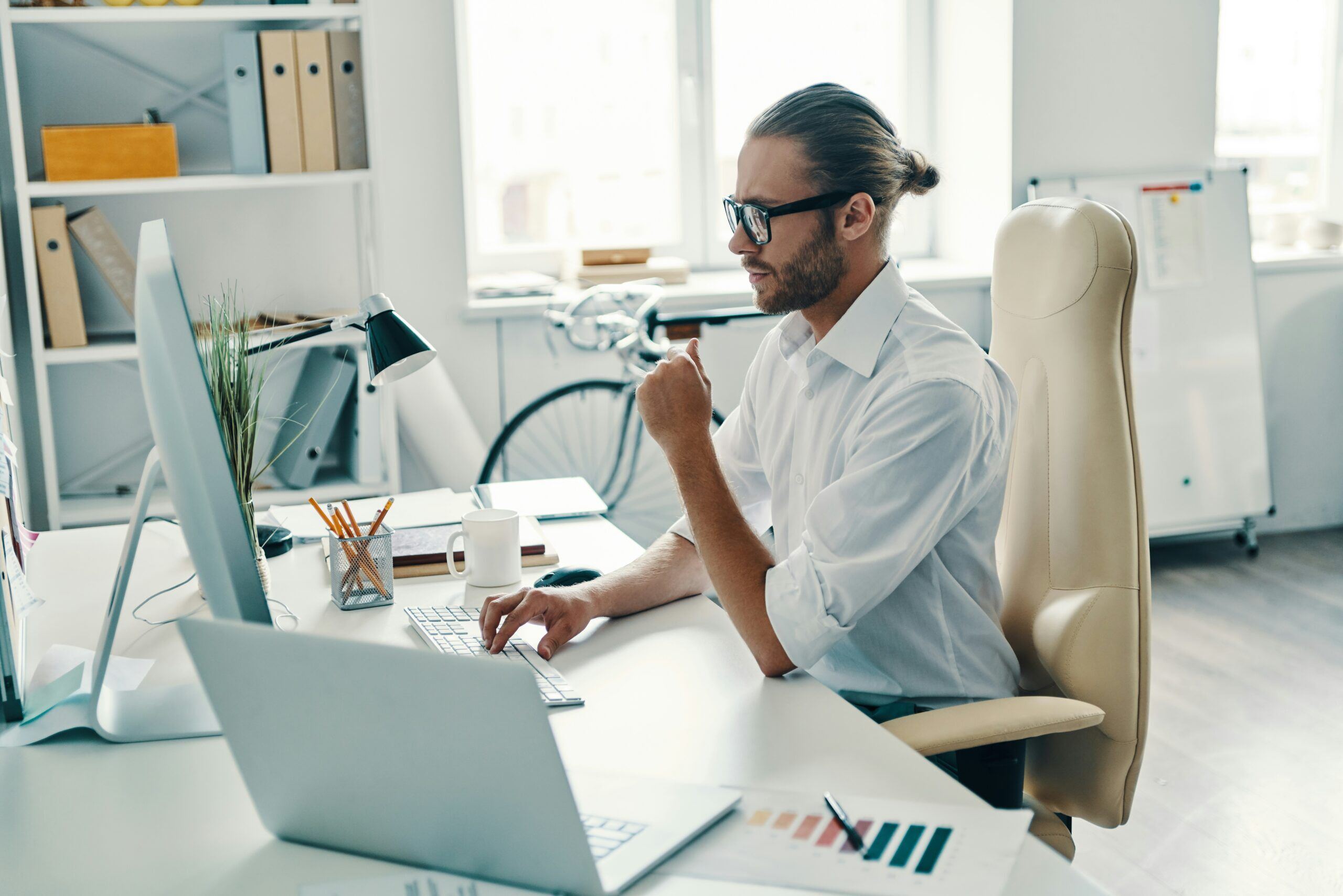 Professional office workspace with analyst at desk, monitor, laptop and printed chart, representing structured SQL Server bottleneck triage and careful technical investigation.